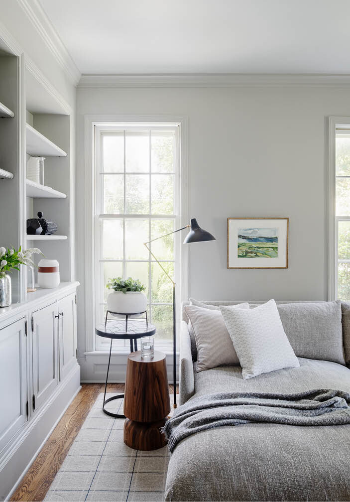 View the family room in a historic Hingham home, highlighting the denim-upholstered chair, light grey sectional, built-ins, and charming historic windows.