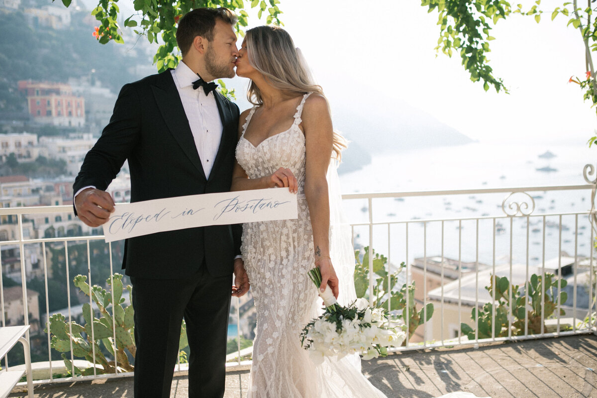 Bride and groom kissing while holding 'Eloped in Positano' sign – stylish destination wedding moment by Studio Rose