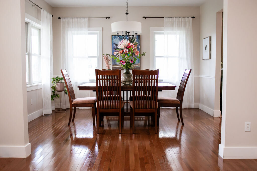 A well-lit dining room featuring a dark brown rectangular dining table surrounded by six wooden chairs with striped cushions. A clear glass vase filled with purple coneflowers and greenery serves as a centerpiece. A large framed painting of a vibrant floral bouquet hangs on the wall behind the table, illuminated by a modern black chandelier with a white drum shade. Natural light fills the room from windows draped with sheer white curtains. The floor is made of polished hardwood, reflecting the light and furniture. A small potted plant stands near one of the windows.