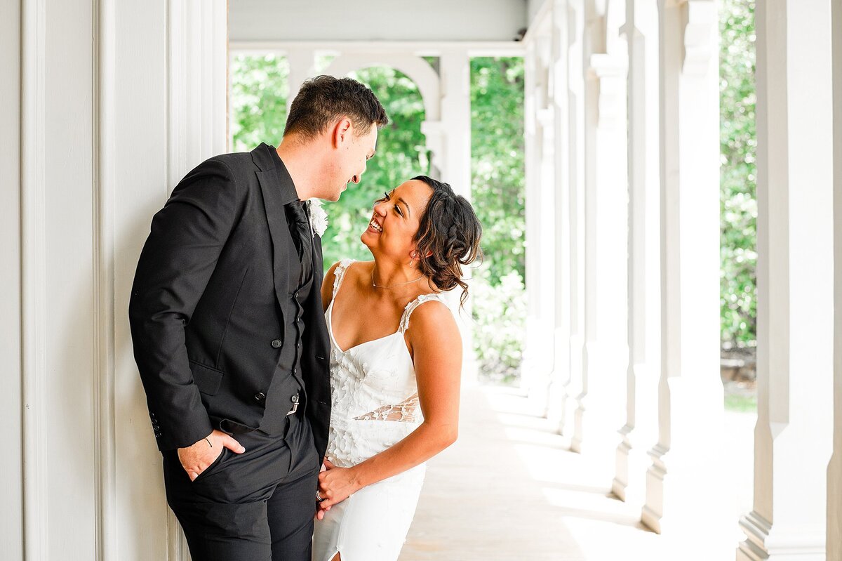 The groom, wearing all black, leans in to kiss the bride, wearing a fitted lace wedding dress with spaghetti straps. They are standing on a white front porch of a historic mansion in Nashville with fluted columns in the background.