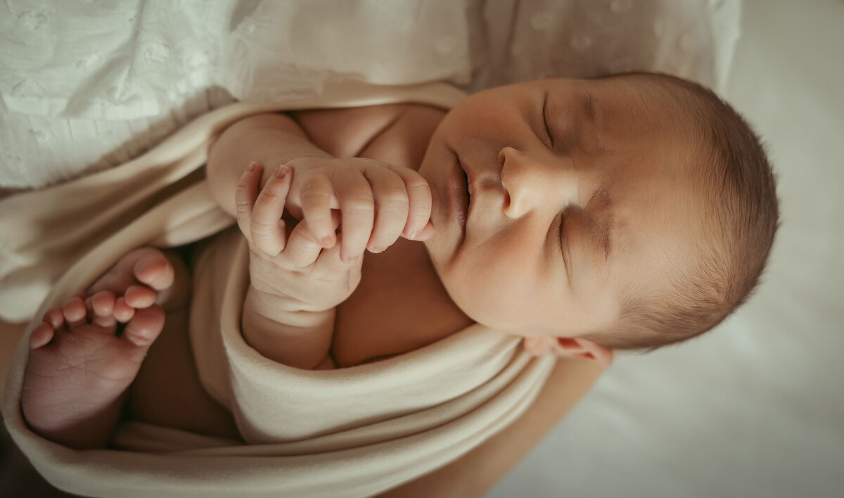 newborn baby sleeping wrapped in a beige blanket holding both his hands together