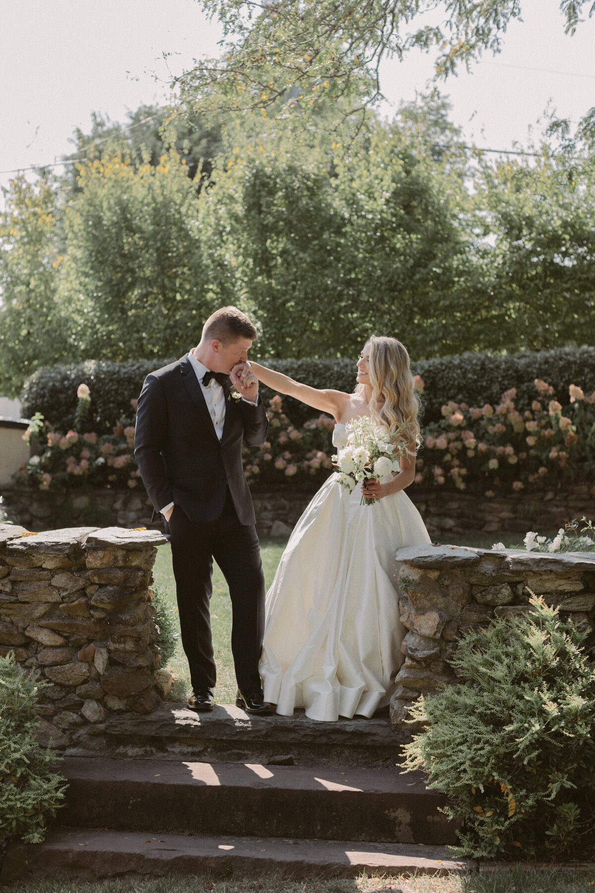 Groom gently kisses the bride’s hand as she smiles, standing together in a sunlit garden surrounded by greenery and blooming flowers.