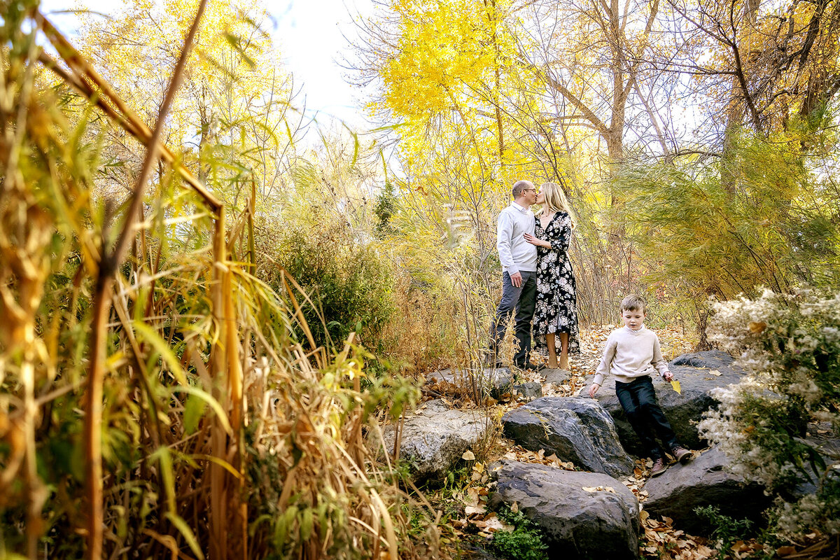 Colorado family photo session in Highlands Ranch in the fall. Boy sits by a creek with his parents close behind and beautiful fall colors surrounding.