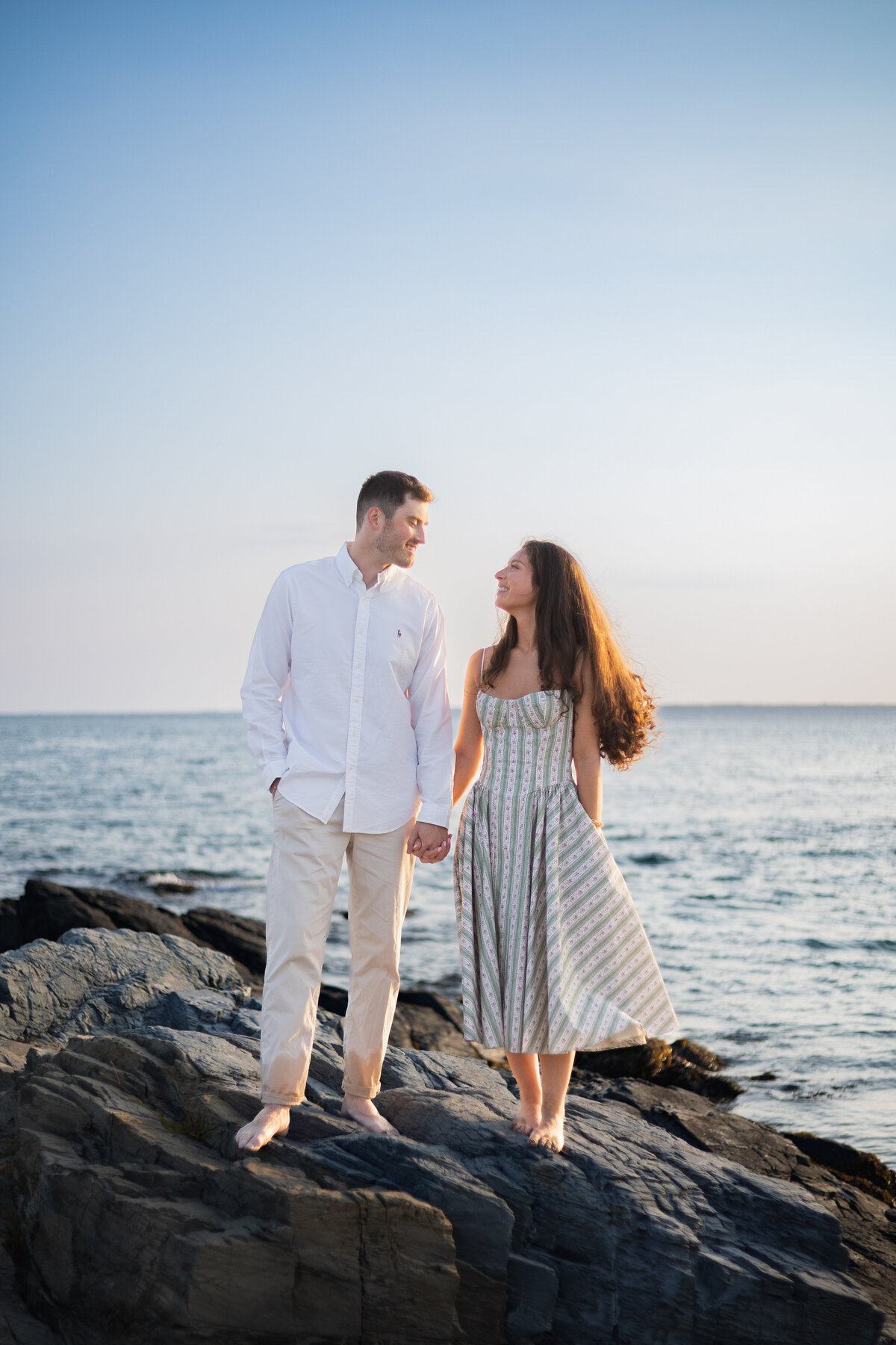 Engagement Photos Rhode Island | Kelsey Sheehan Photography Timeless Rhode Island Weddings | A couple stands barefoot on rocks by the sea, holding hands and smiling at each other. The sky is clear, setting a serene and romantic tone.
