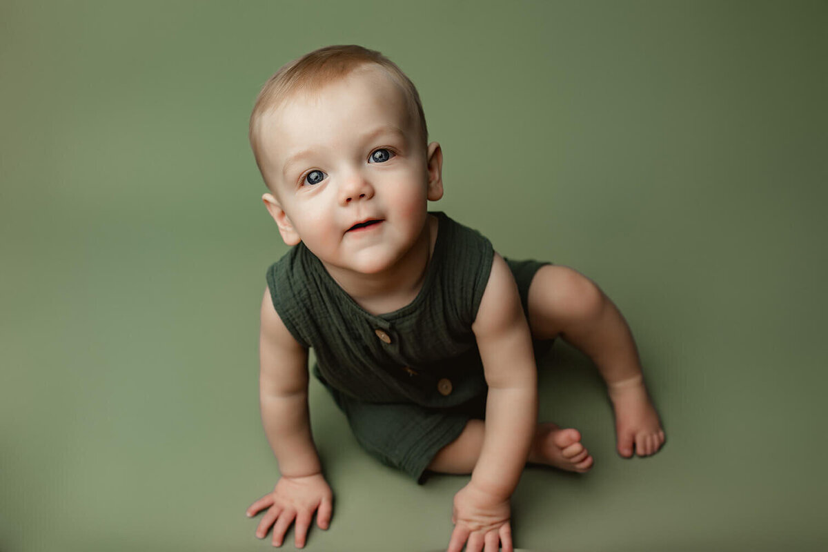 A boy sits on a green drop wearing a green onesie while looking at the camera