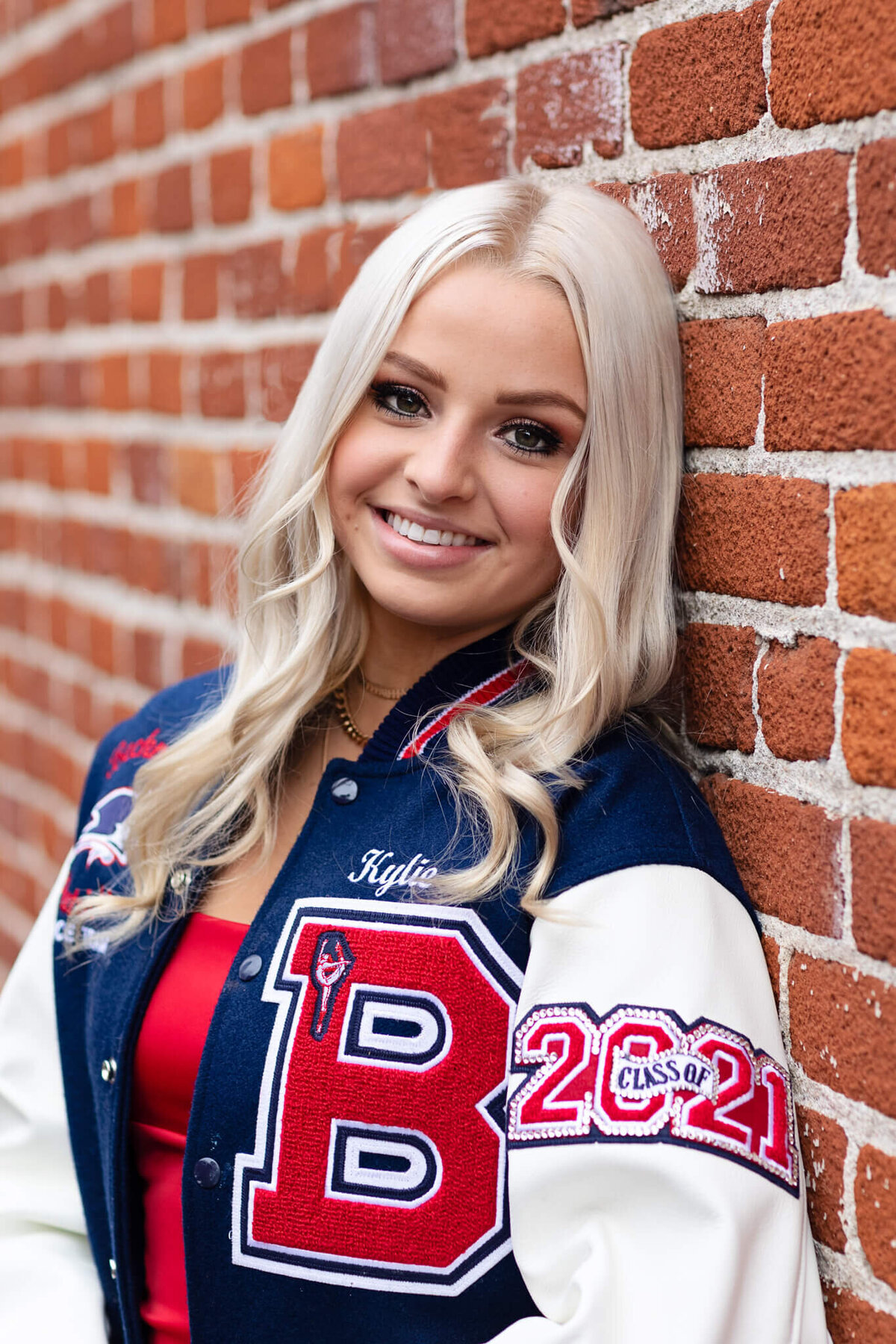 A young woman with long blonde hair is leaning against a brick wall, smiling. She is wearing a blue and white varsity jacket with the letter B and Class of 2021 on it.