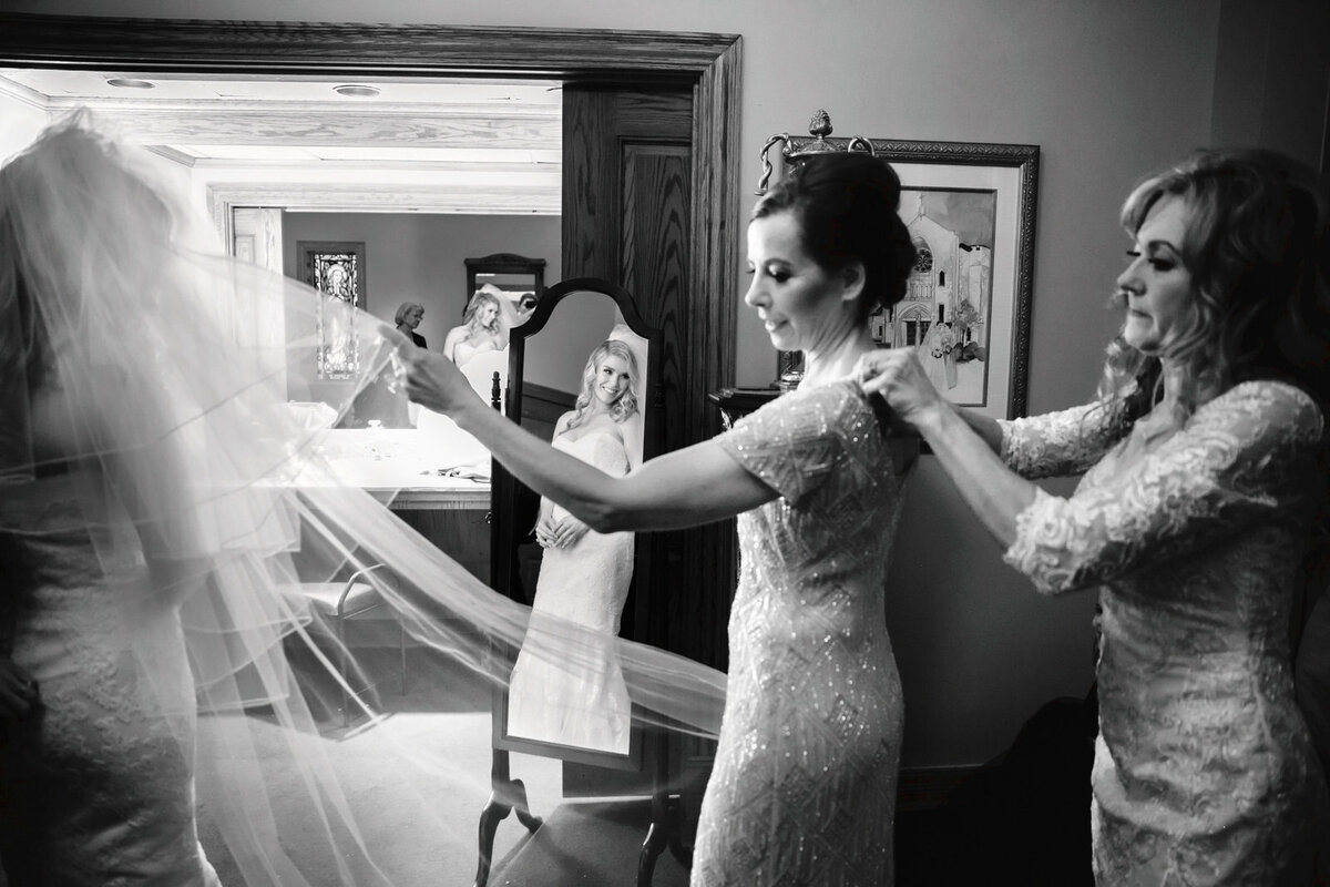 Candid moment at St. Thomas Aquinas Church in Dallas with the bride getting ready while her mother adjusts her dress and the mother of the groom helps the mother of the bride