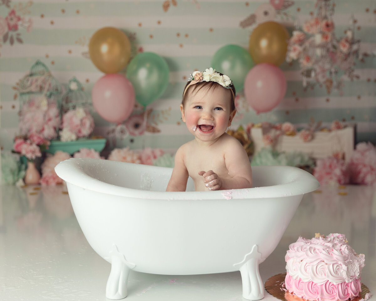 A toddler girl wearing only a headband sits in a bathtub during her birthday photo session