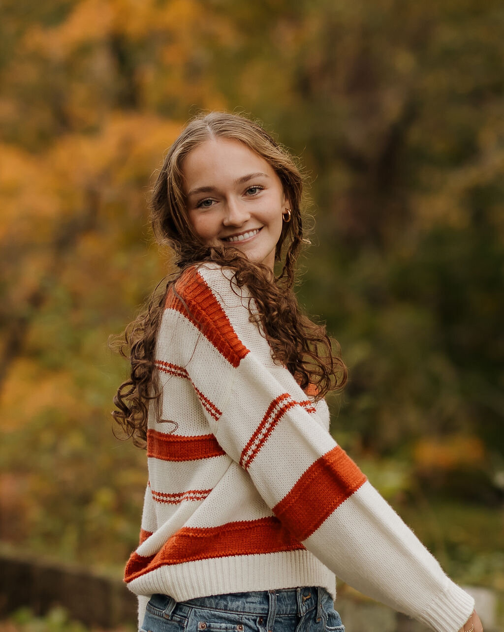 A young woman with long curly hair smiles while sporting a white sweater with red stripes and jeans. Captured by a New England couple photographer, she stands outdoors against a blurred backdrop of autumn trees, embodying the region's portrait photography charm.