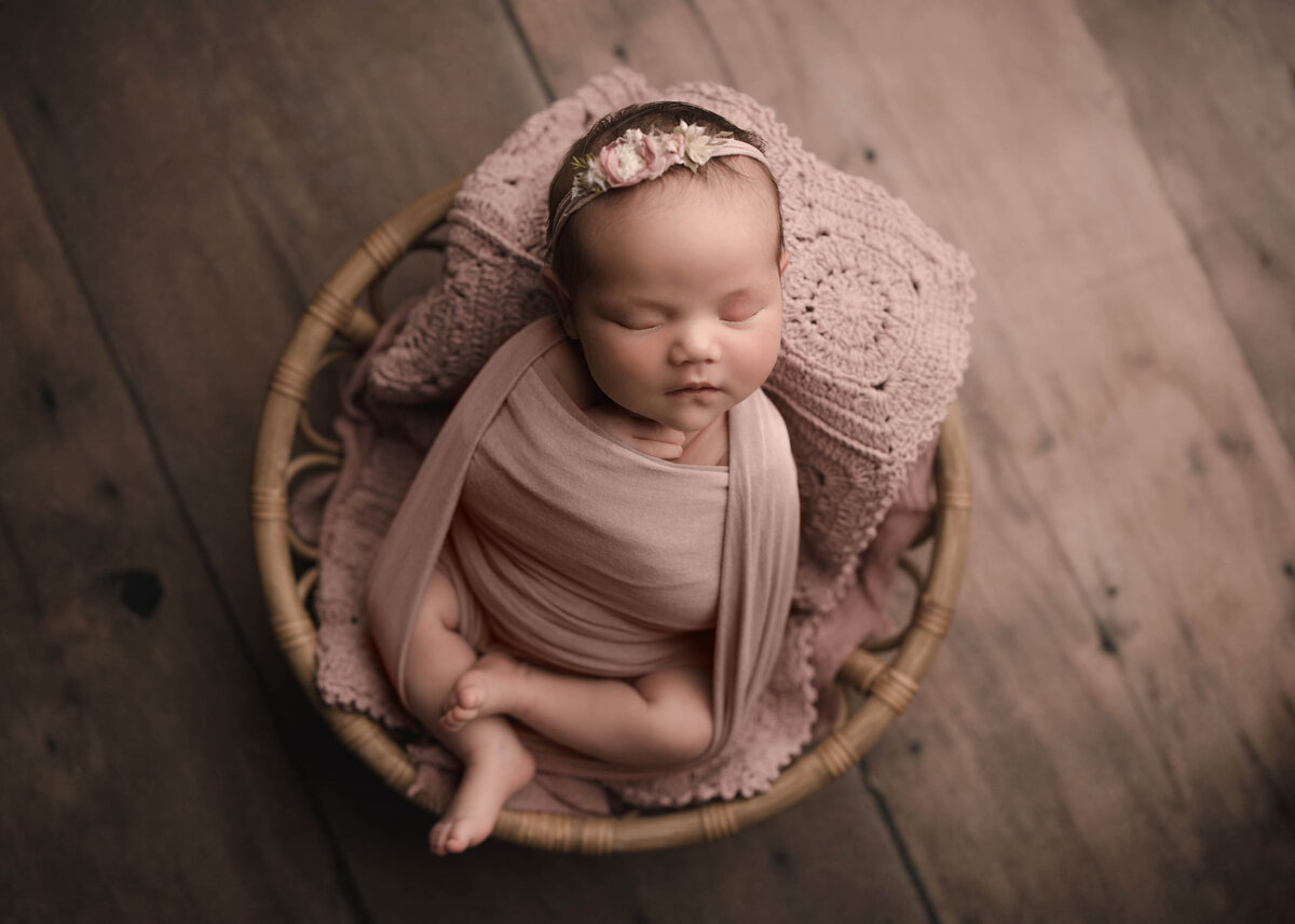 Aerial image. Baby girl is wrapped in a blush stretch wrap with her legs out and folded atop of her. Sh e is sleeping in a basket and wearing a delicate floral headband. Captured by Menifee newborn photographer Bonny Lynn Photography