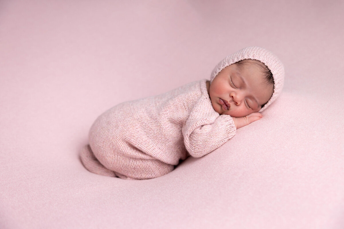 newborn baby wearing a pink knitted romper and hat asleep on pink fabric