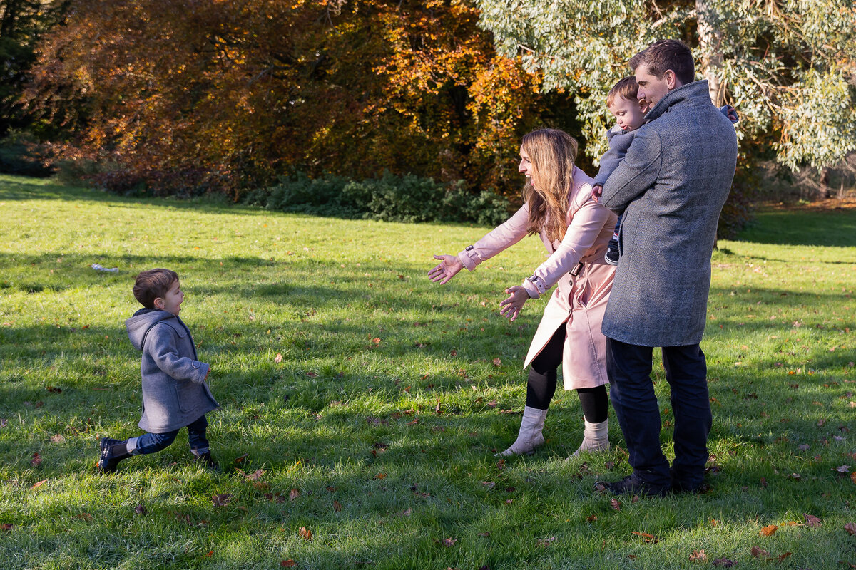 A family of four enjoys a sunny day in the park; a woman reaches towards a young boy, with another child and a man beside her, surrounded by autumn trees.