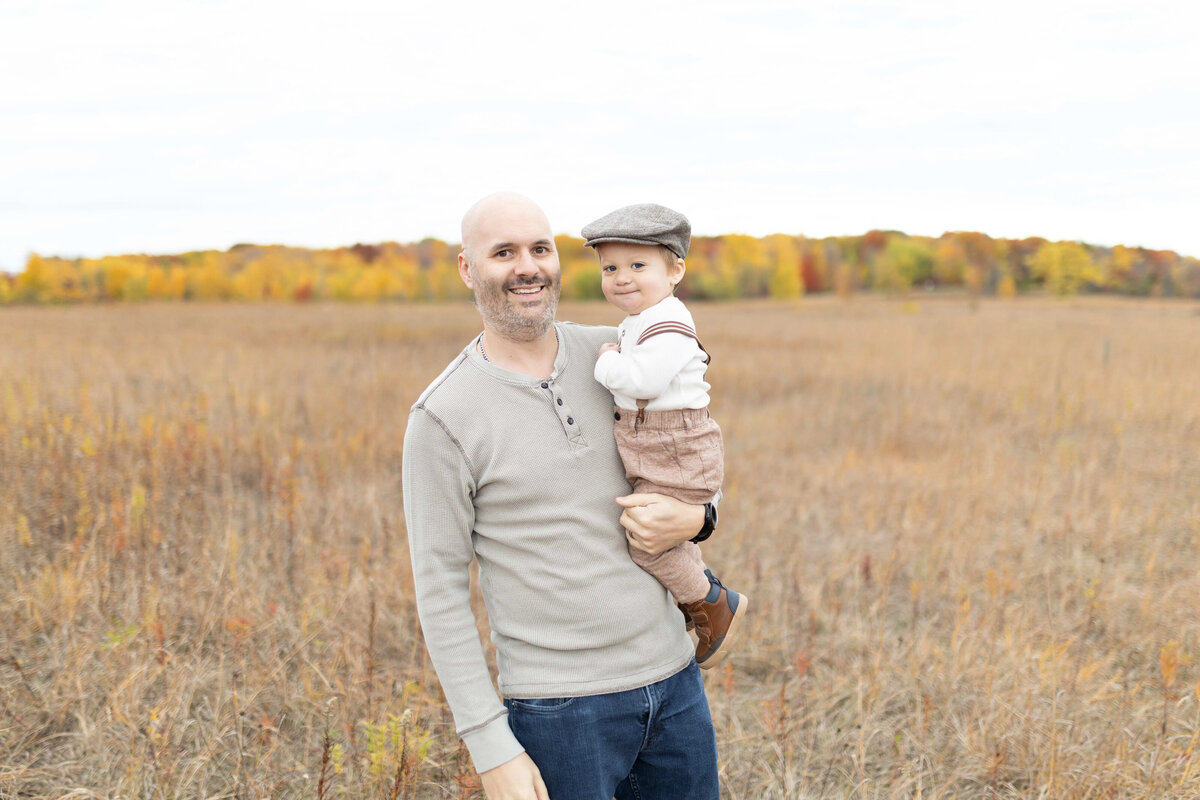 Dad holding toddler boy in brown suspenders i n fall field - outdoor family session Maple Grove
