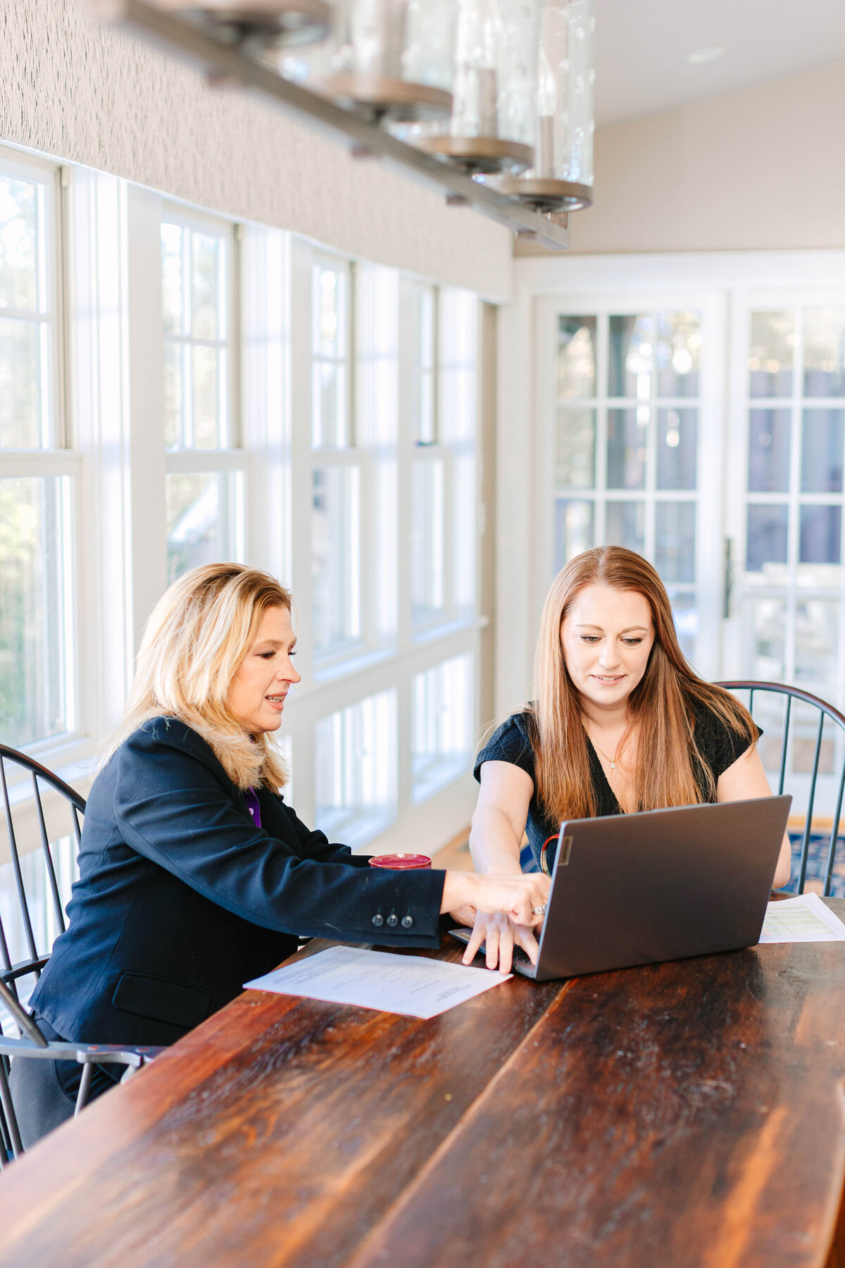 Two women sit at a rustic wooden table in a bright, sunlit room with large windows. One woman, wearing a black blazer, gestures toward a laptop screen while the other, in a black top, listens attentively. Papers and a red drink lid are scattered on the table, suggesting a collaborative work session