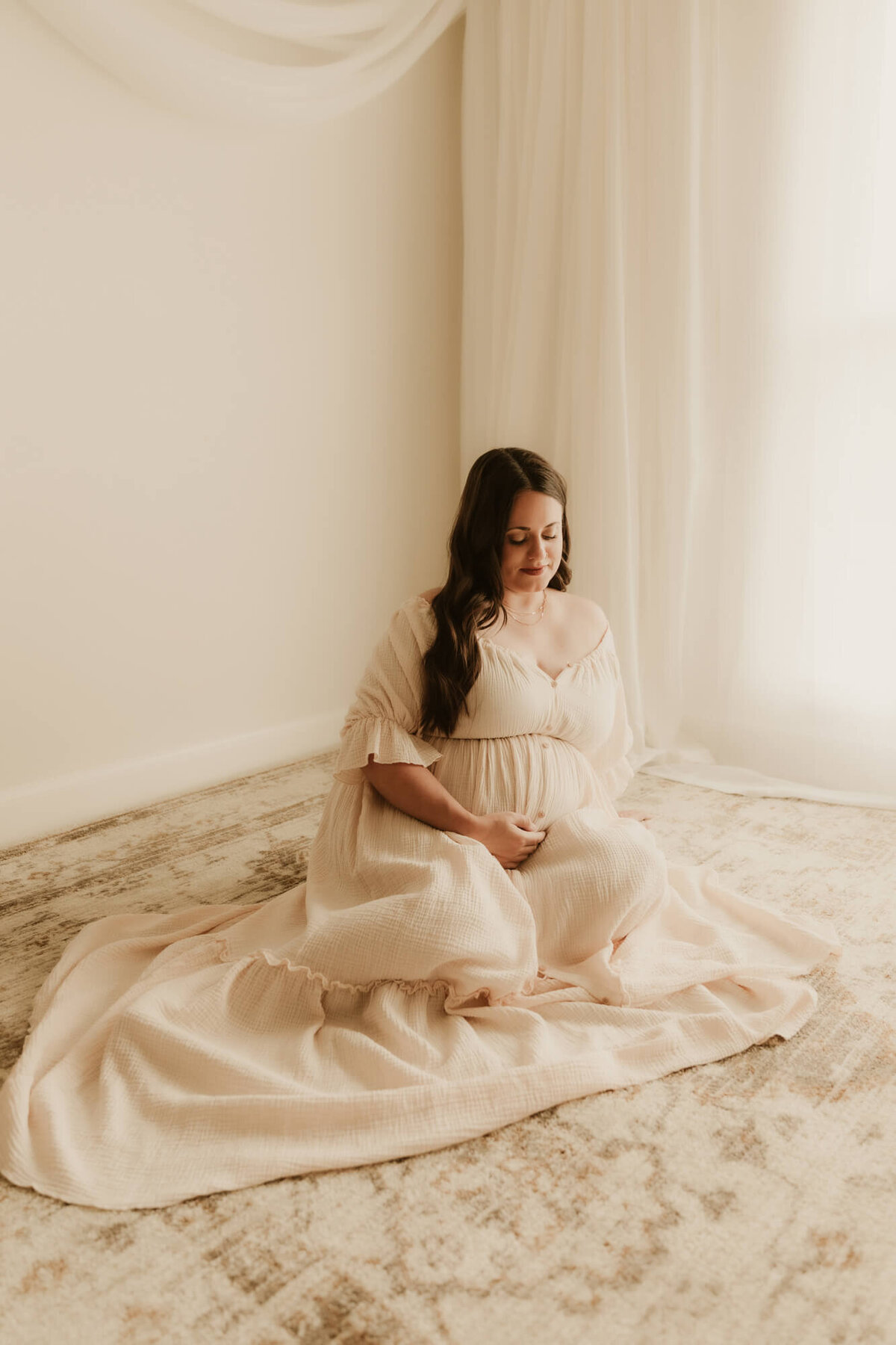 Mother looks at her baby bump while sitting down on a rug and holding her belly.
