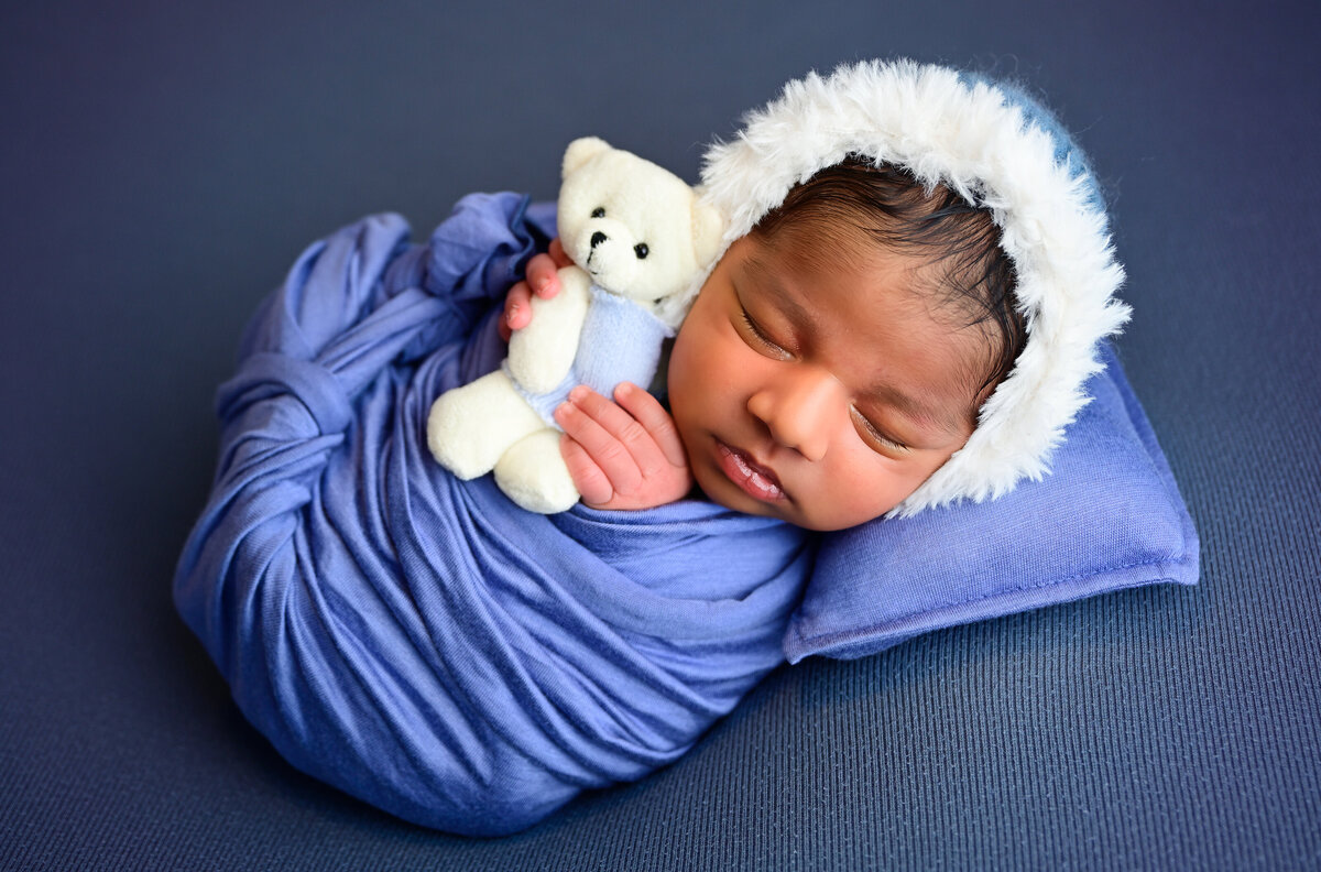 Kitsap newborn photo session with baby wrapped in blue, wearing a blue bonnet with fur trim, sleeping holding a lovie teddy bear.  Image captured by Beth Benecki of E. Benecki Photography.