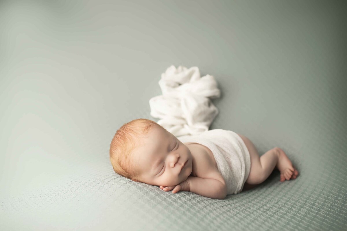 Tiny red-haired newborn boy in a classic newborn pose, resting his head on his hands on a neutral-toned backdrop