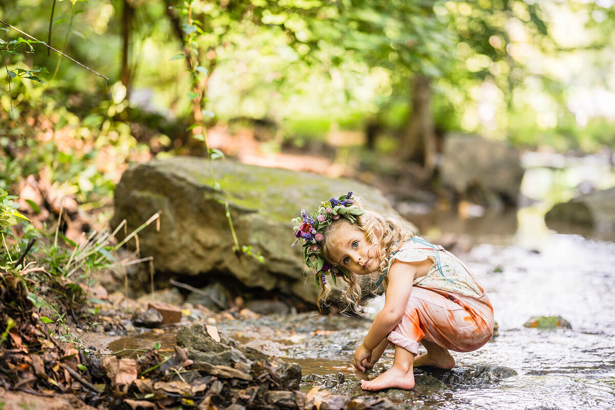 A child wearing a pair of dyed overalls and a flower crown is found mid-squat playing in the water and peeks over her shoulder as a photo of her is taken in the creek at Fishburn Park.