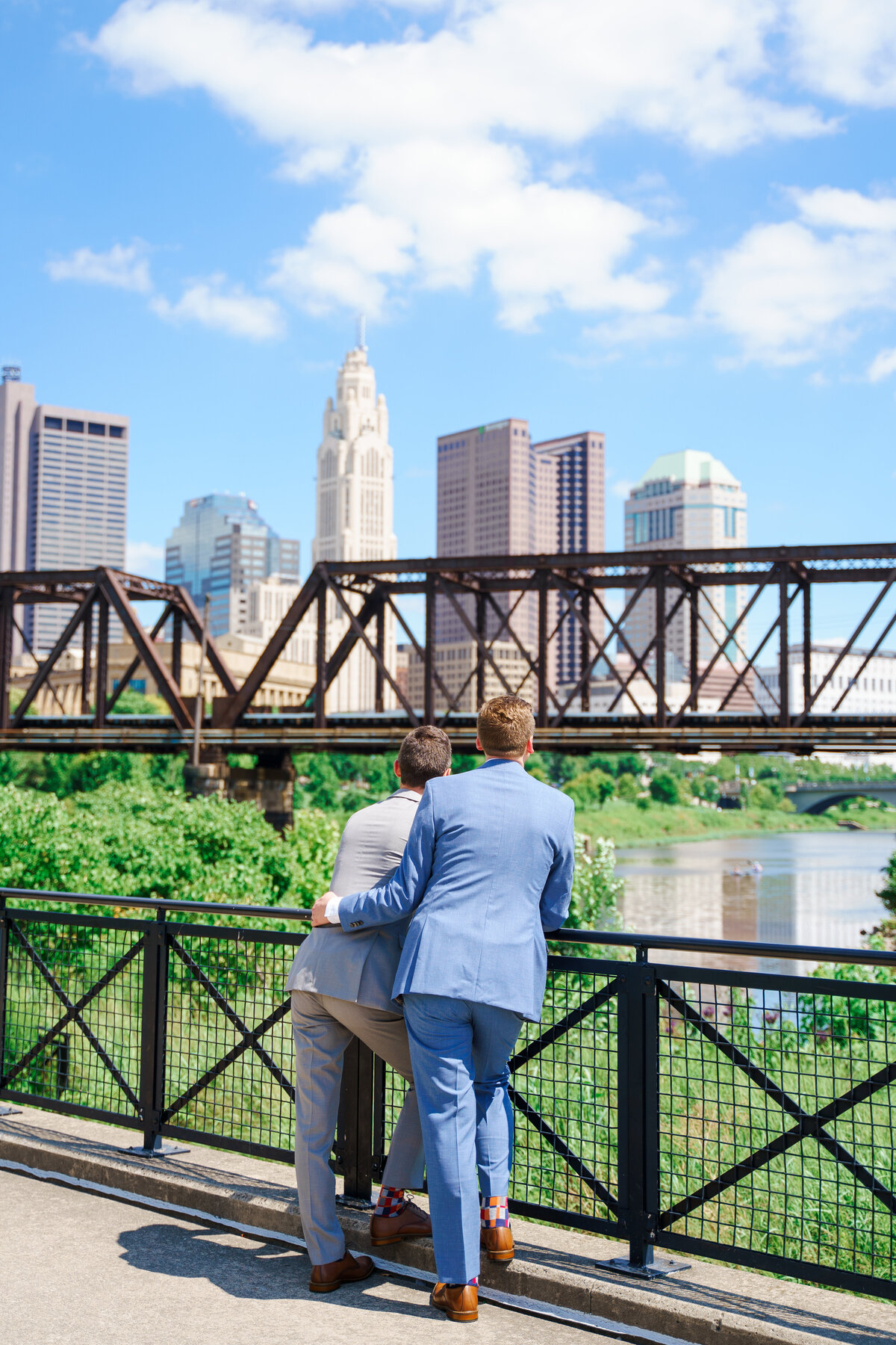 A groom puts his arm around his groom as they look off into the Columbus skyline at their wedding at North Bank Park Pavilion.