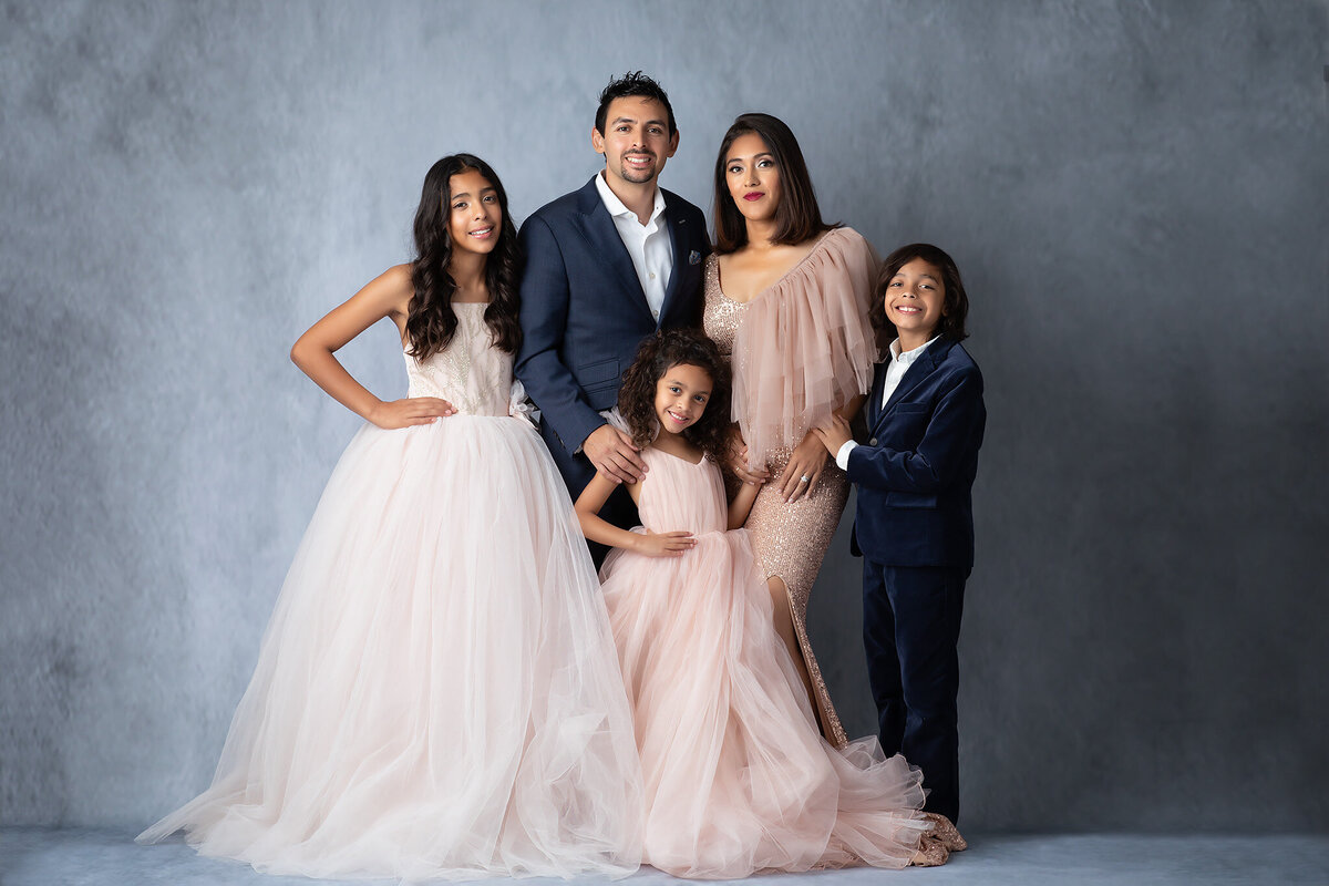 Dallas family photographer capturing a family portrait in a studio with soft lighting. The family of five poses together in elegant attire, including tulle gowns and velvet suits, set against a gray backdrop for a timeless family photograph.