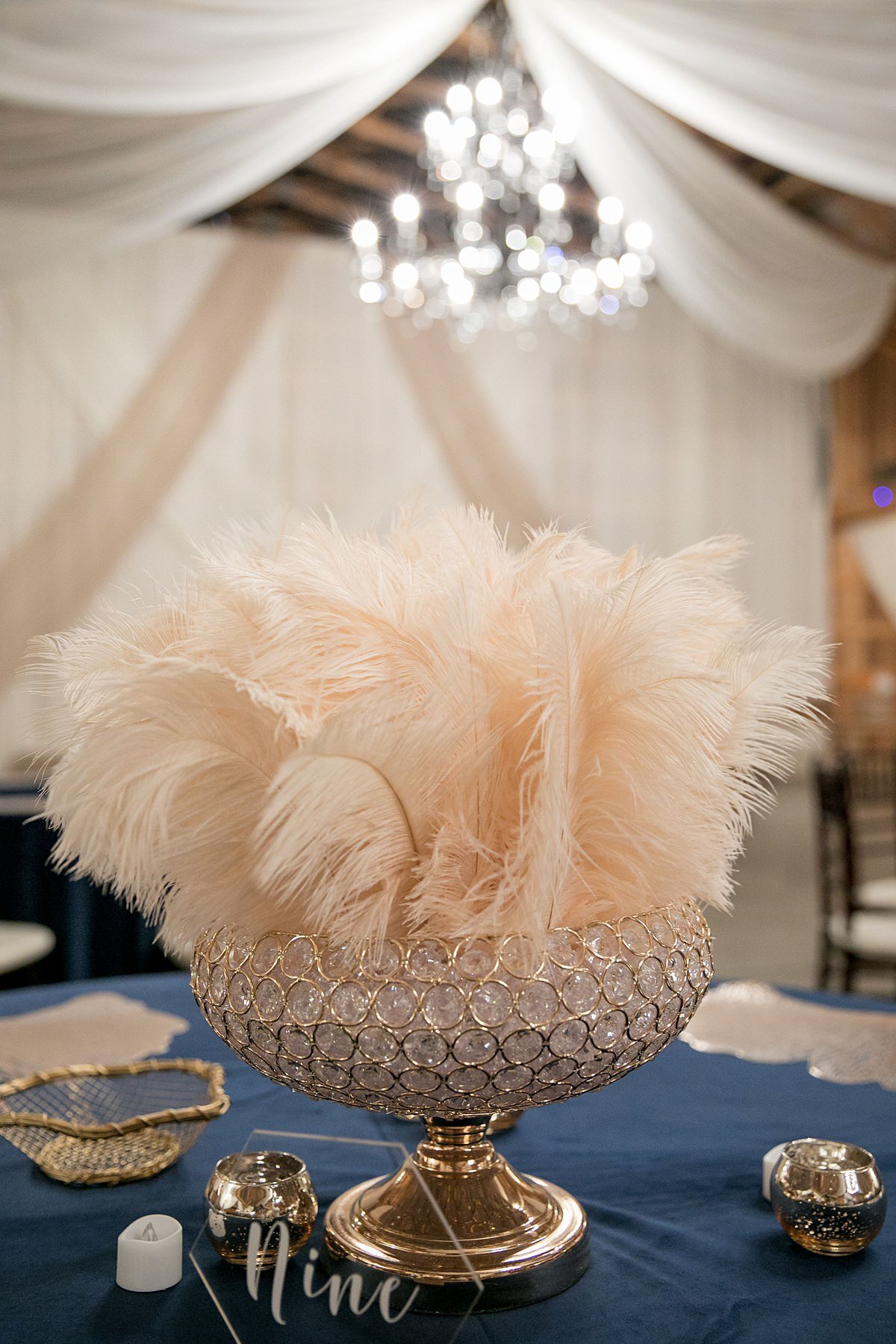Large gold and crystal footed bowl with large plume of ivory ostrich feather centerpiece on a navy blue table cloth with an acrylic geometric table number with a gold bowl of Hershey's kisses and gold votive candles. The ceiling is draped with white and ivory sheer fabric accenting the gold and crystal candelabra chandelier which matches the white and champagne drapery on the back wall.