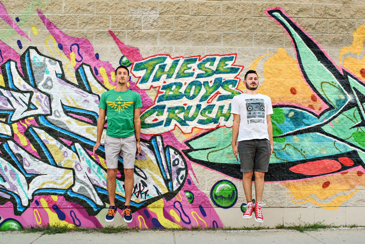 A same sex couple jump in front of a wall of graffiti in chigago that says, these boys crush.