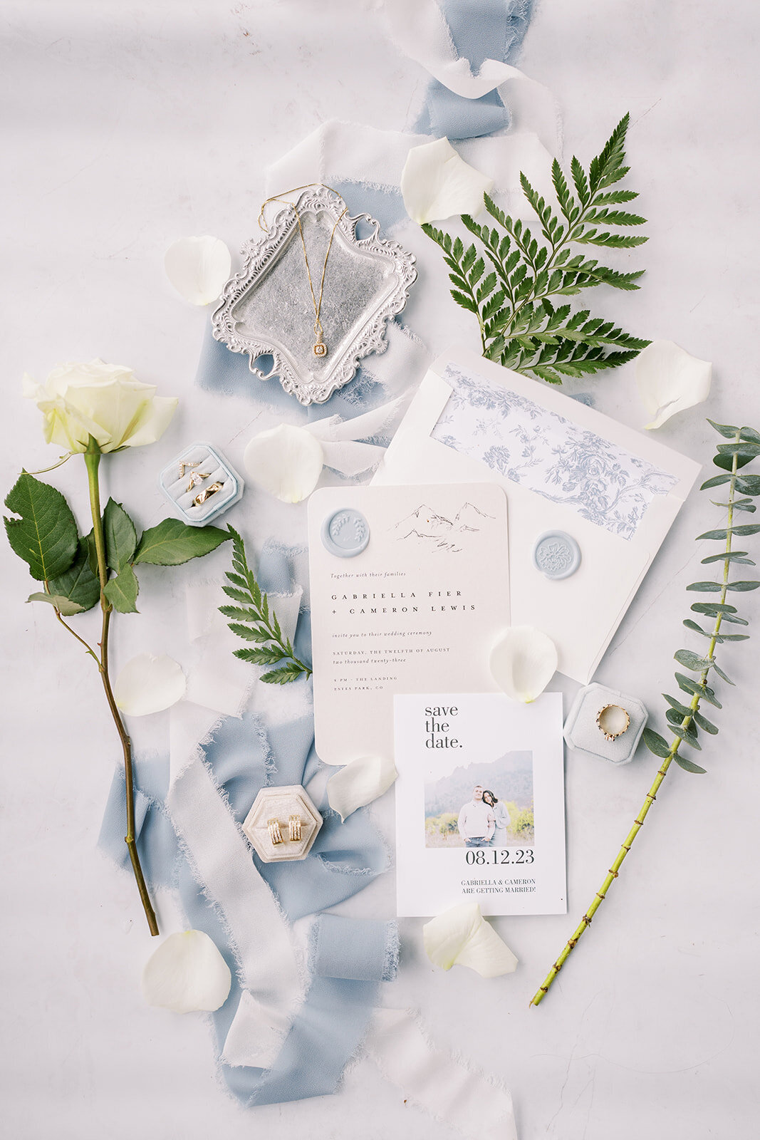 Wedding detail flat lay with light blue and white invitation suite and wedding rings.