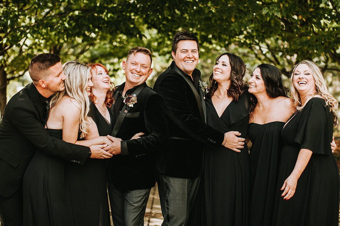 Two grooms wearing black tuxedos share smiles outside with their wedding party also wearing all black.