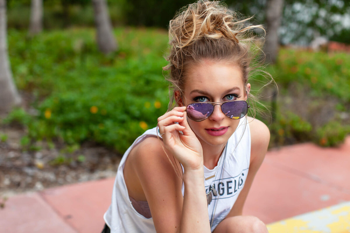 A close up portrait of a high school senior girl looking over her sunglasses with her hair piled on top of her head in a messy bun. Photograph by Dynae Levingston