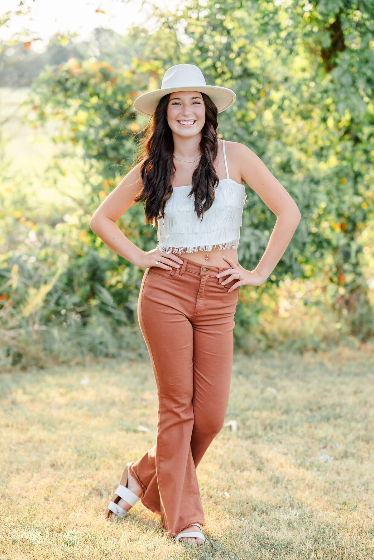 A high school senior, wearing orange pants, a fringe top, and white hat poses with her hands on her hips and a big smile on her face. The sun peeks through some bushes behind her. Photo by Justine Renee Photography at Bells Mill Park in Chesapeake, Virginia.