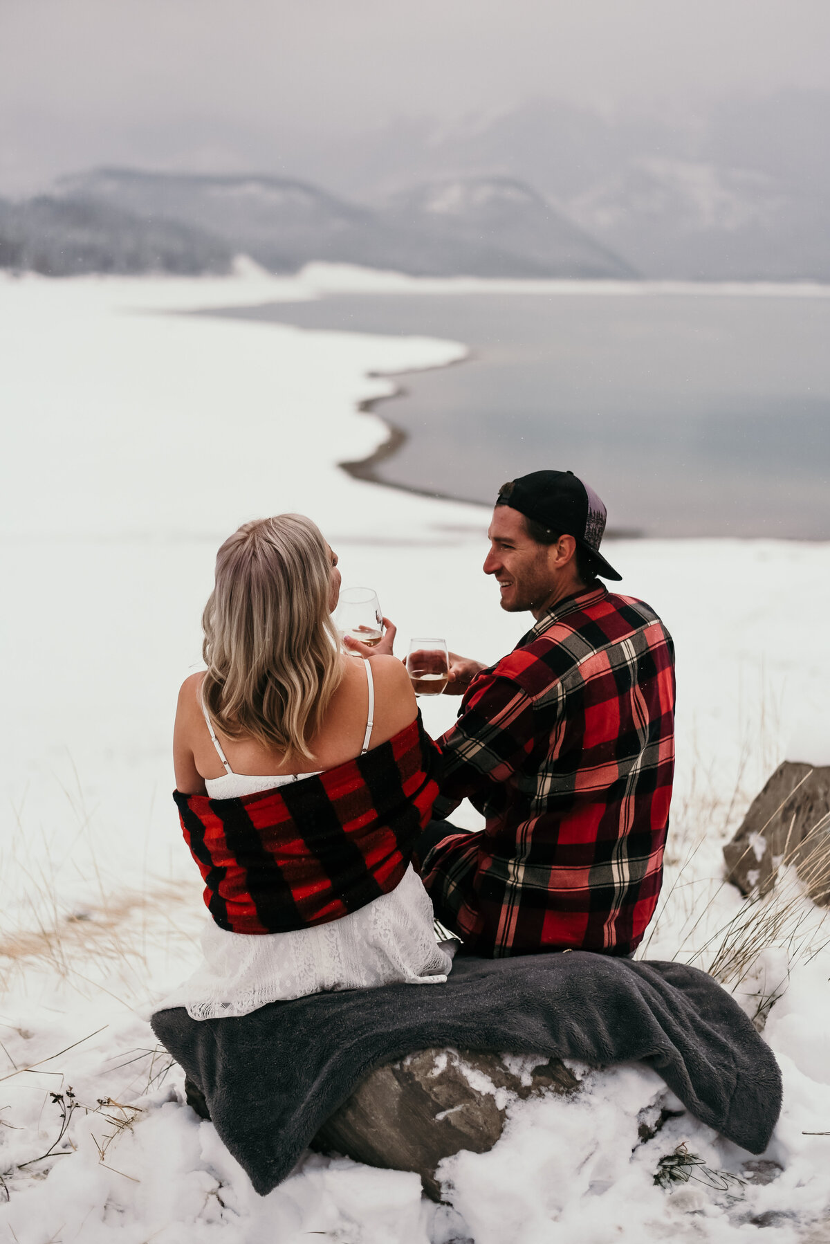 Man and woman share a drink with snow surrounding them looking onto mountain and lake views