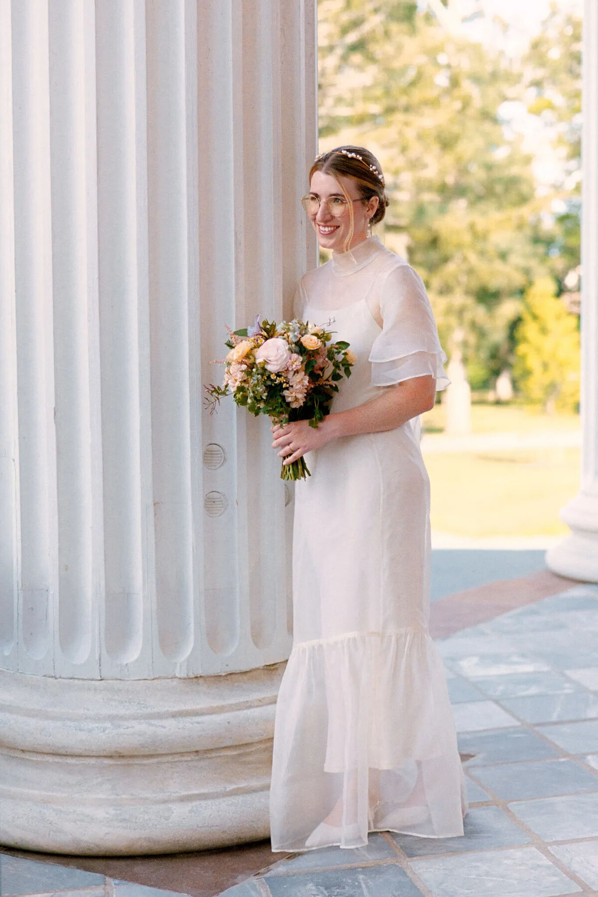 A person in a white wedding dress holding a bouquet of flowers stands smiling beside a large white column outdoors, with greenery and sunlight in the background—captured beautifully by a film photographer NJ.