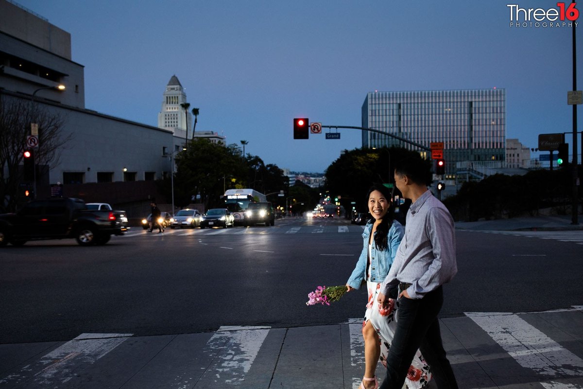Engaged couple walk across a sidewalk at night in Downtown Los Angeles during engagement photo shoot