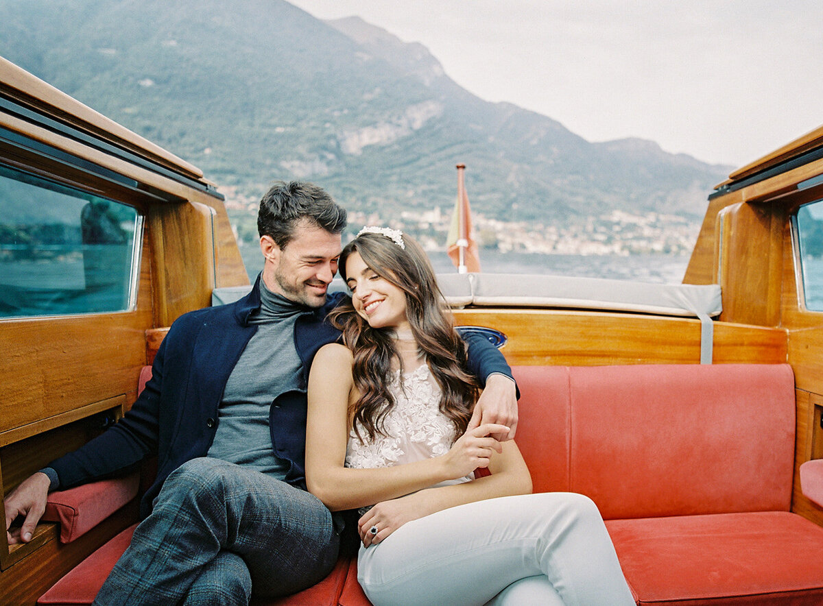 Film photograph of bride and groom on a wooden boat on Lake Como in Italy she is wearing a white halter jumpsuit with her hair down in loose waves and he is wearing grey and navy cashmere sweater and coat photographed by Italy wedding photographer