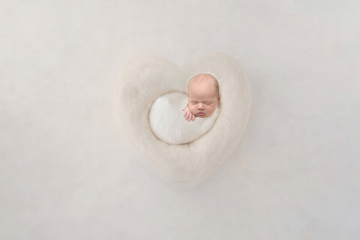 baby wrapped in blanket  thats white while sitting in a heart shaped basket and white backdrop