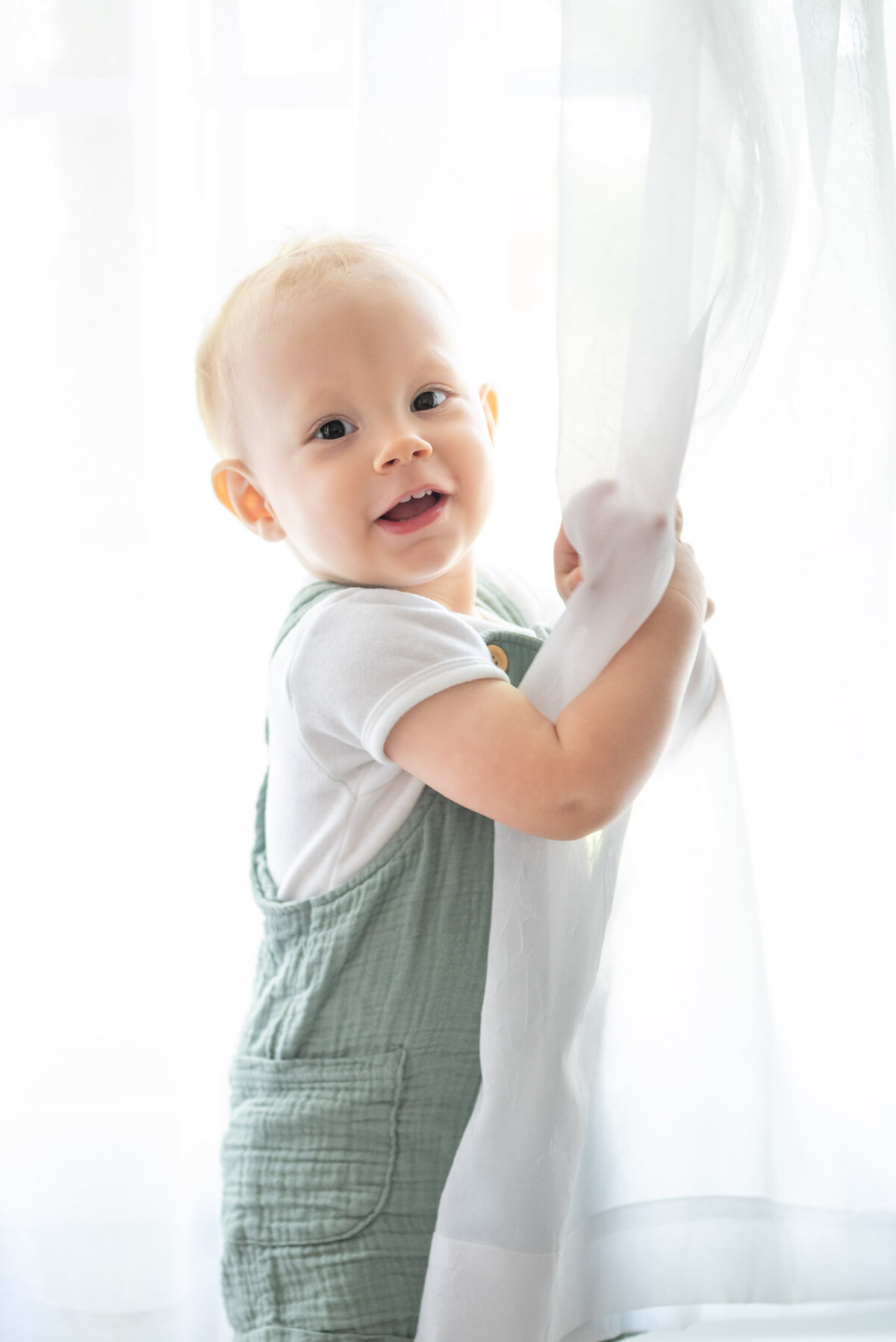 A baby boy peeks around  a curtain  in a photography studio in Huntsville Alabama