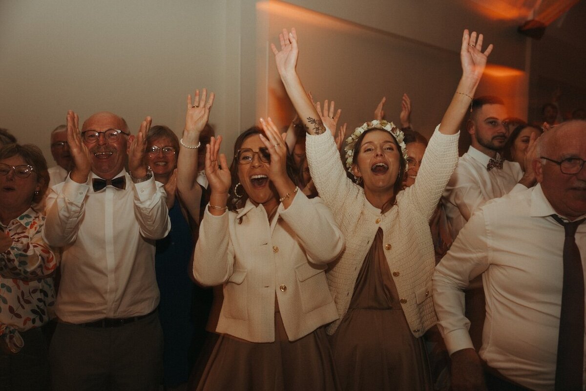 Un groupe d'invités en liesse, les bras levés, applaudissant avec enthousiasme pendant la soirée du mariage. Reportage de mariage en Bretagne par Kamescop.