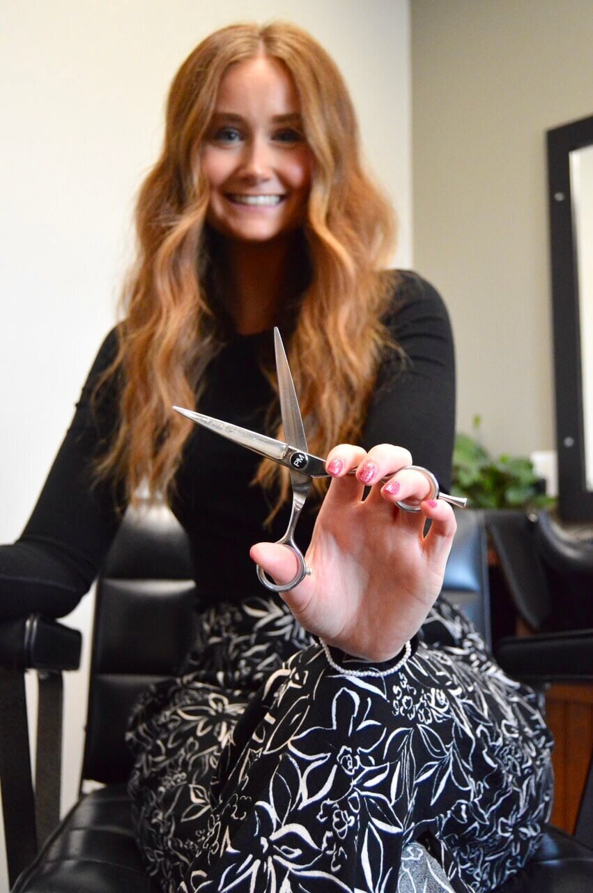 A smiling hairstylist holding a pair of scissors, seated in a salon chair, showcasing the friendly and professional service at 212 Salon, Spa, & Barbershop.