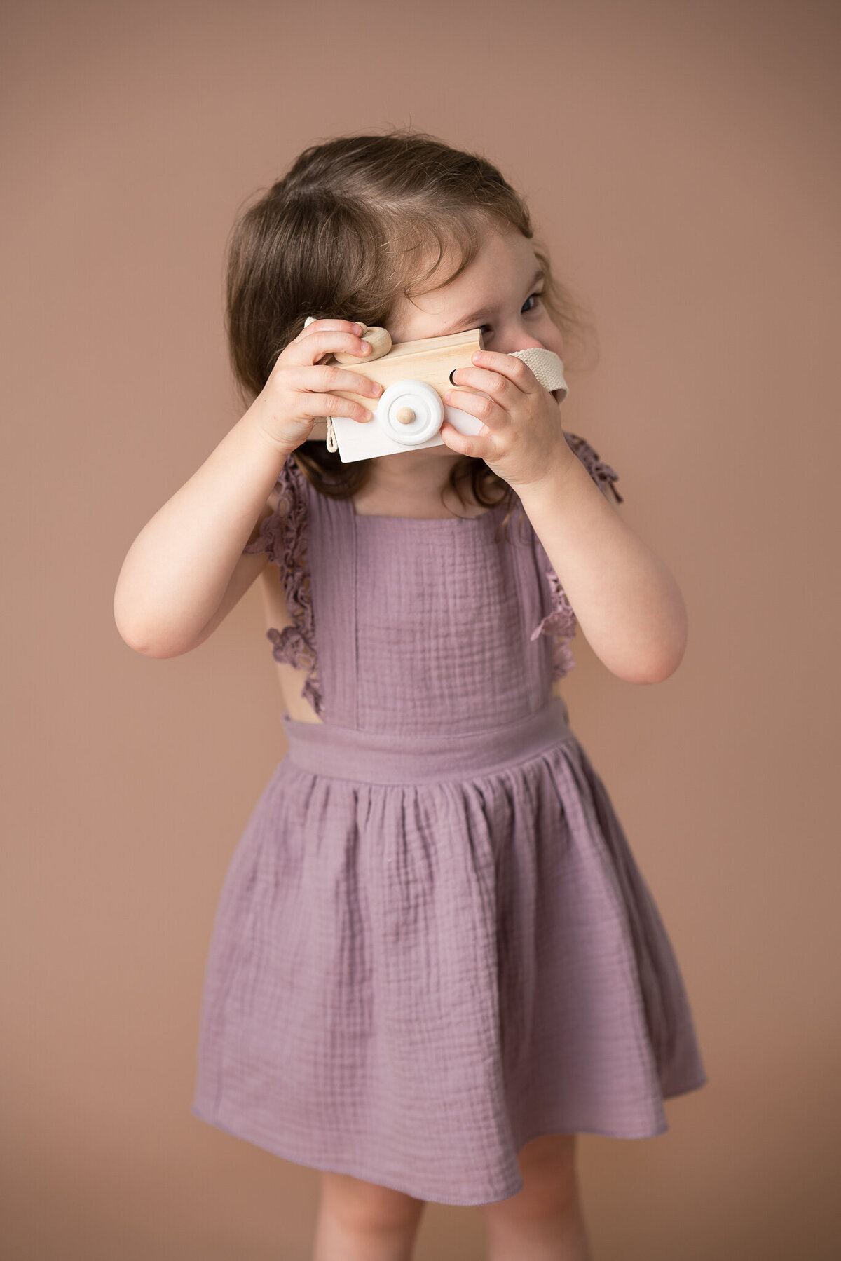 A young girl in a purple dress is holding a toy wooden camera.