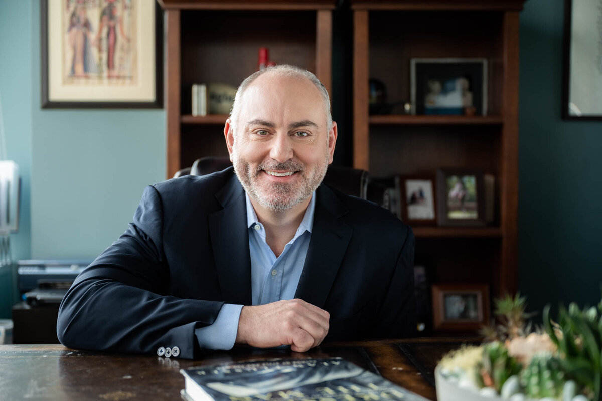 a man seating behind a desk with his arm on the desk smiling to the camera