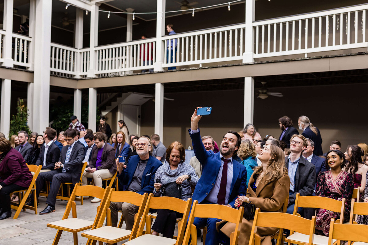 A group of people sit on yellow chairs at an outdoor wedding. A man in a blue suit holds a phone up, smiling and taking a selfie. Other attendees around him chat, smile, or look towards the front.