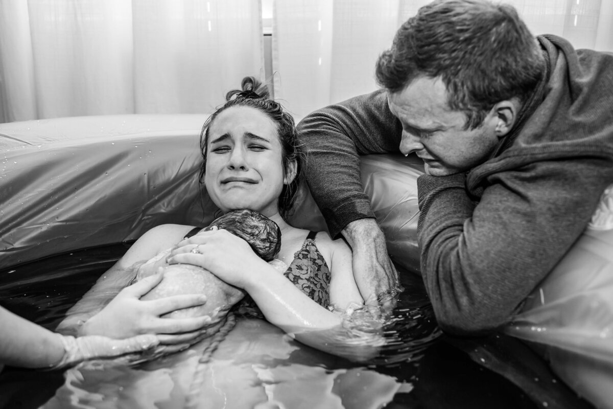 black and white image of a crying mom holding her newborn in a birthing pool while her husband cries over her shoulder