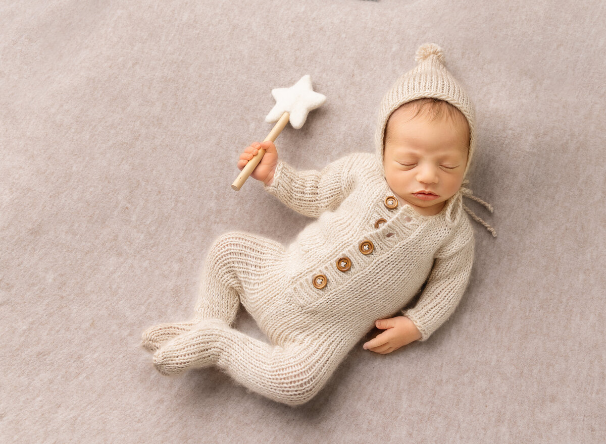 Baby boy sleeping on is back for a brooklyn newborn photoshoot. Baby is in a cream knit onesie and holding a felt magic wand. Captured by top Brooklyn newborn photographer Chaya Bornstein