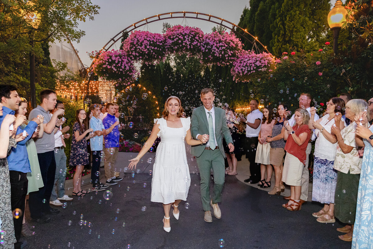 The bride and groom joyfully walk hand in hand through a path lined with guests blowing bubbles, under an archway adorned with pink flowers and twinkling lights at Le Jardin in Sandy, Utah.