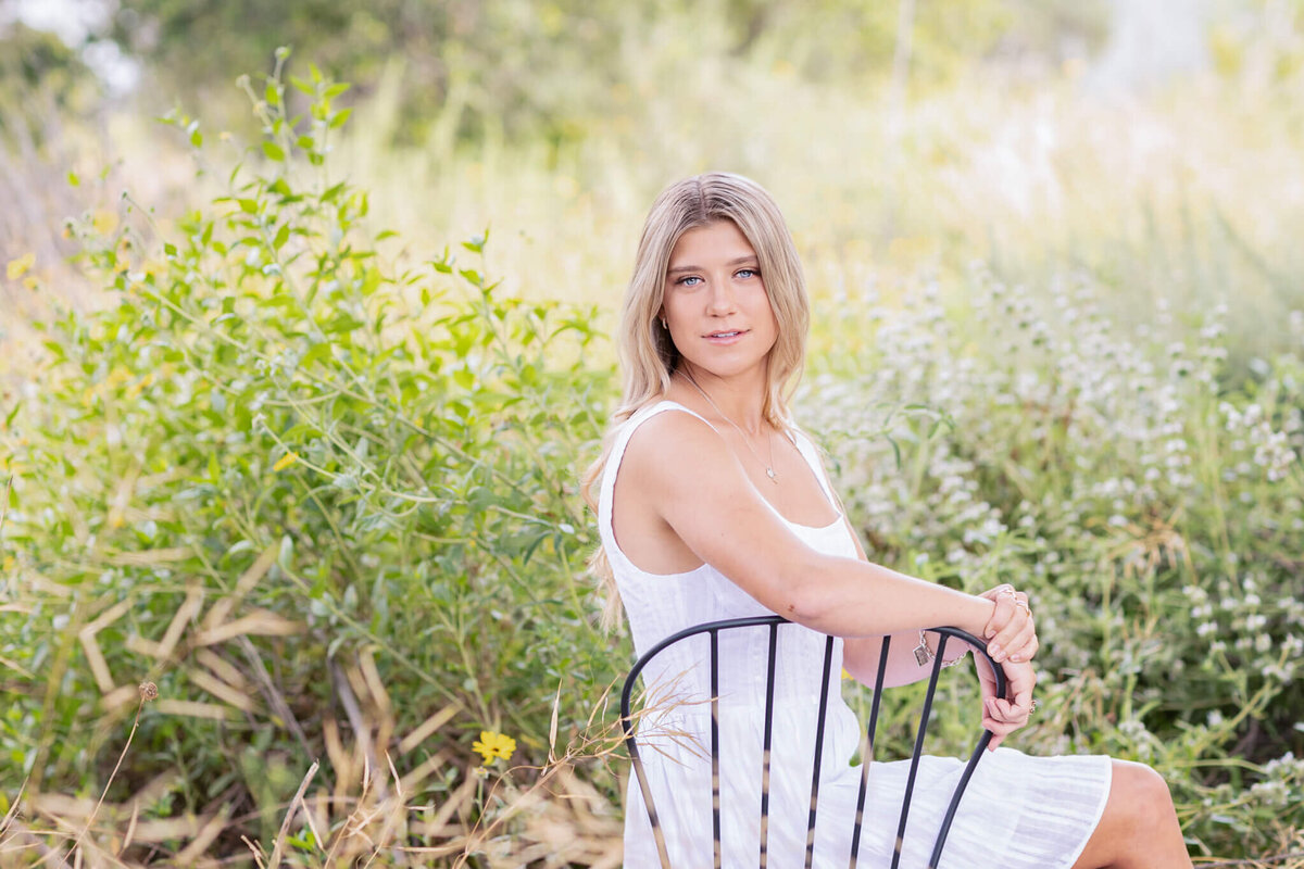 A person with blonde hair sits on a black chair in a field. They are wearing a white dress and looking directly at the camera. The background is filled with green foliage and tall grass, creating a serene outdoor setting.