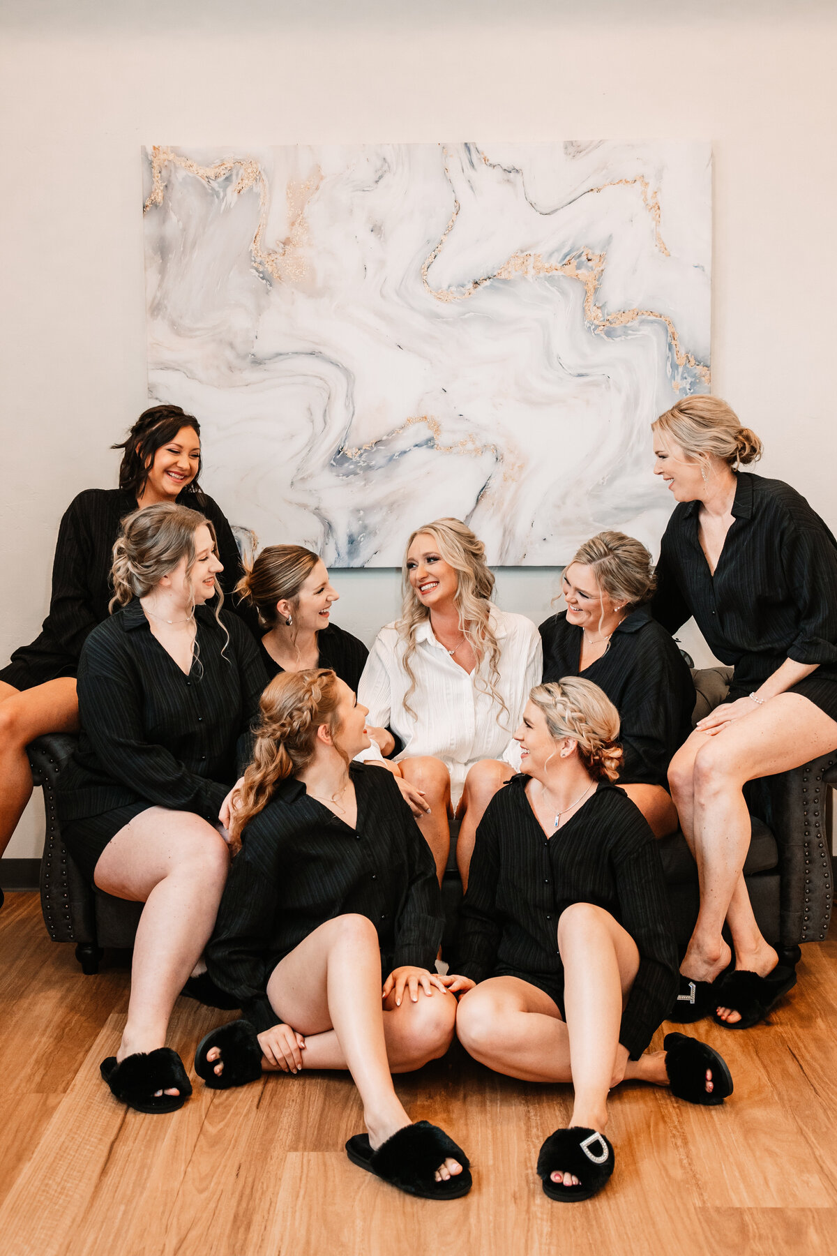 A bride in white pajamas sits in the center of a couch with her many bridesmaids in black pajamas sitting around her