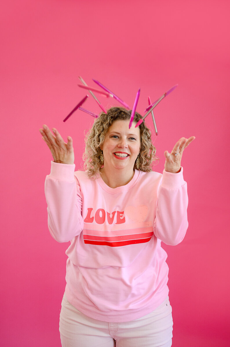 A branding photographer and brand photographer takes a picture of a woman business owner wearing all pink in front of a pink background throwing pink pens in the air during her personal branding photo session with a personal branding photographer during a brand photography session.