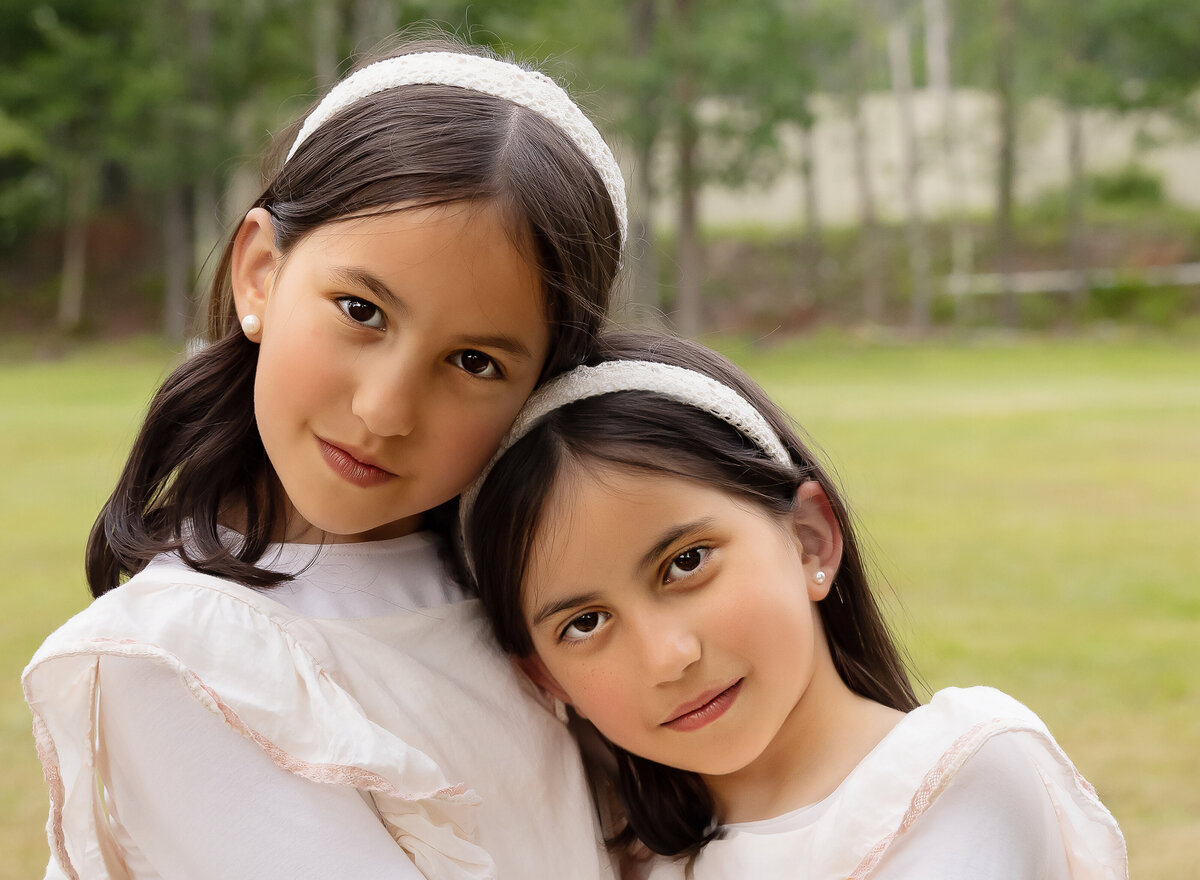 Two sisters in white with white headbands are captured in a brooklyn, ny park for a family photoshoot. Both girls have a serious expression and their heads are touching. Captured by brooklyn family photographer Chaya Bornstein.
