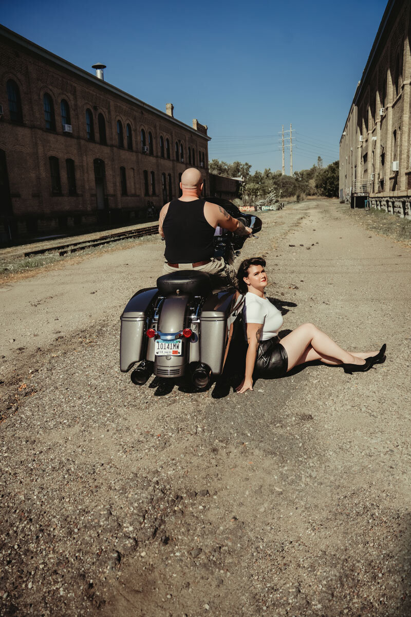 black motorcycle with a bald man sitting on it and a woman with pinup shorts and white shirt sitting on the ground by the motorcycle in Minneapolis