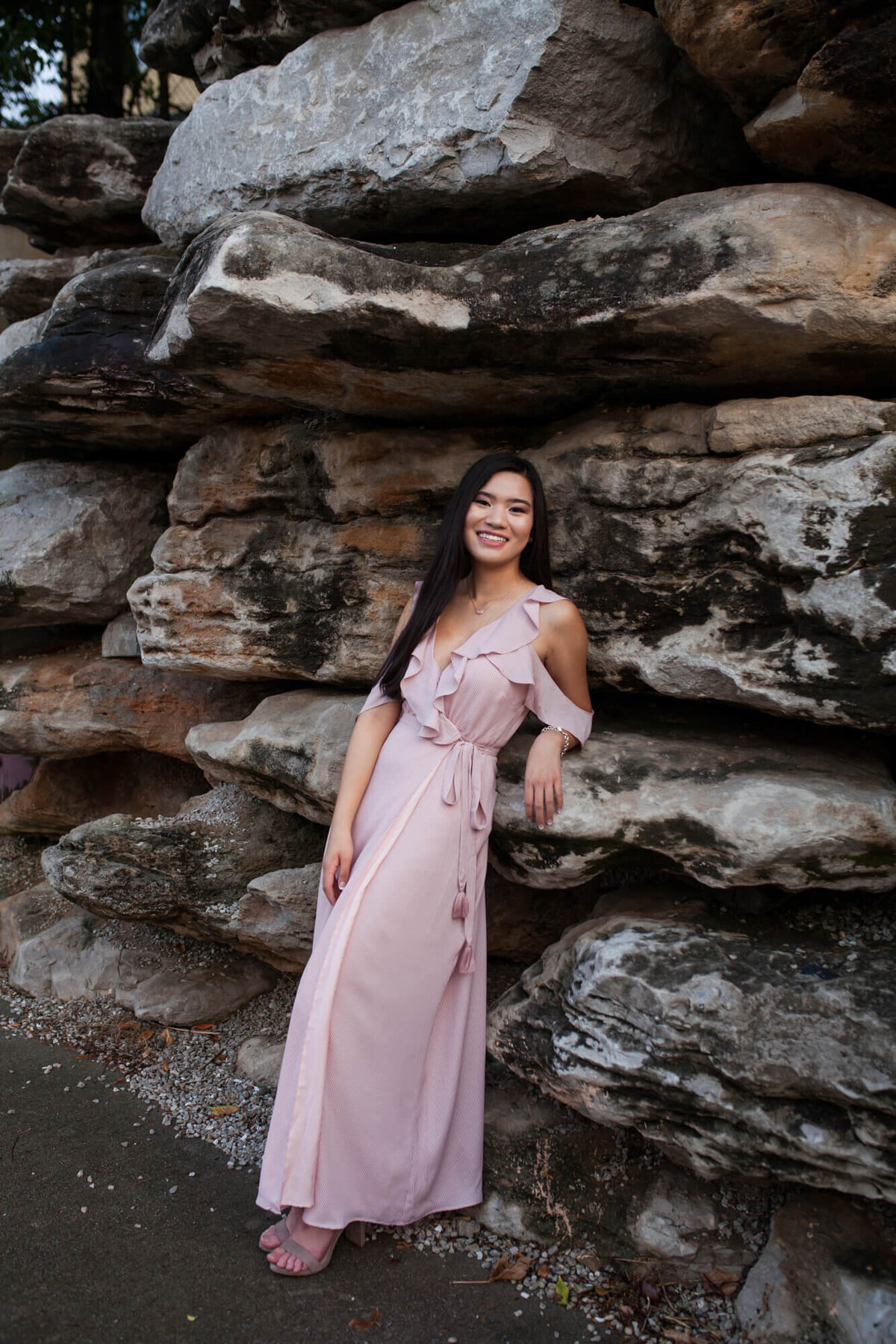 A lovely dark haired teen girl in a light pink dress poses for her senior pictures in front of a rock wall. Captured by Springfield, MO senior photographer Dynae Levingston.